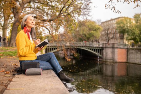Téléchargez les photos : Inspiration. Femme assise sur la rive de la rivière, prenant des notes et regardant inspiré - en image libre de droit
