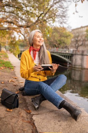Téléchargez les photos : Inspiration. Femme assise sur la rive de la rivière, prenant des notes et regardant inspiré - en image libre de droit