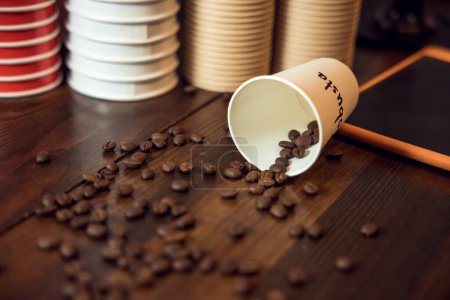 Photo for Close-up of an overturned disposable paper cup lying on the tablet computer and robusta beans scattered on the tabletop - Royalty Free Image