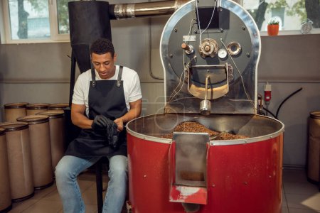 Photo for Calm focused young worker sitting beside the coffee roasting machine and putting on nitrile gloves - Royalty Free Image
