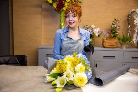 Foto de En una florería. Linda joven pelirroja de pie a la mesa en una floristería - Imagen libre de derechos