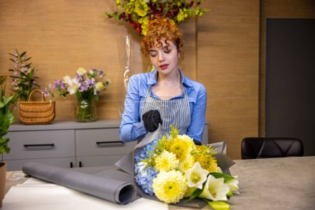 Foto de En una florería. Linda joven pelirroja de pie a la mesa en una floristería - Imagen libre de derechos
