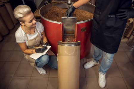 Téléchargez les photos : Maître rôti regardant le collègue verser des grains de café torréfiés de la goulotte de libération dans le récipient - en image libre de droit