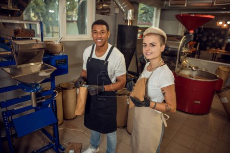 Foto de Sonrientes tostadores de café jóvenes satisfechos sosteniendo bolsas de café de papel y de pie ante una máquina de sellado - Imagen libre de derechos