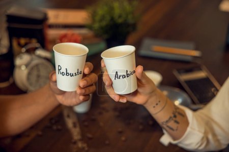 Foto de Foto recortada de dos personas con tazas de café de papel desechables en sus manos sentadas a la mesa - Imagen libre de derechos