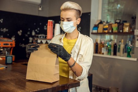 Photo for Concentrated coffee shop worker in the face mask packing the paper bag for a customer at the bar counter - Royalty Free Image