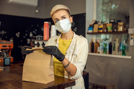 Téléchargez les photos : Employé de café réfléchi dans un masque jetable emballant le sac en papier au comptoir du bar - en image libre de droit