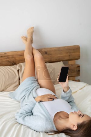 Foto de En línea. Mujer joven embarazada acostada en la cama con las piernas arriba y navegar por Internet - Imagen libre de derechos