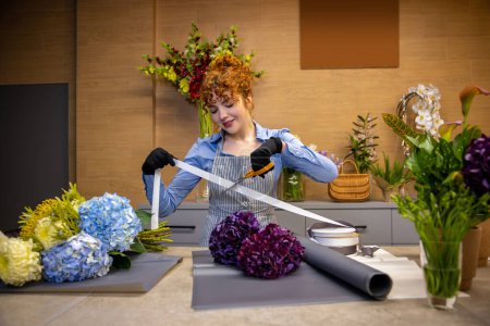 Foto de Florista en el trabajo. Linda florista de jengibre mirando involucrado mientras que hace una cubierta para el ramo - Imagen libre de derechos
