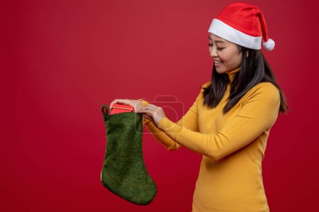 Photo for Christmas gift. Smiling and excited young woman in santa hat putting gifts into stocking - Royalty Free Image