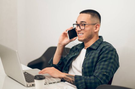 Téléchargez les photos : Joyeux jeune travailleur de bureau asiatique dans les lunettes assis à l'ordinateur portable communiquant sur le téléphone portable - en image libre de droit