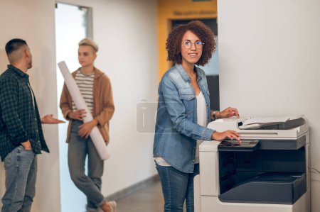Photo for Smiling joyous young African American female office worker pressing the button on the copy machine and looking ahead - Royalty Free Image