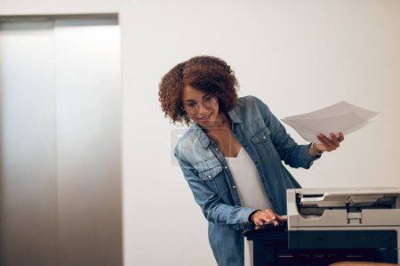Téléchargez les photos : Sourire heureux jeune employé de l'entreprise afro-américaine dans les lunettes en appuyant sur le bouton sur le photocopieur - en image libre de droit