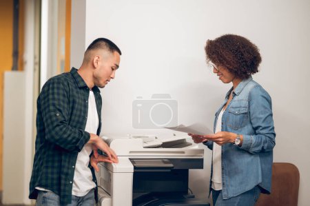 Photo for Smiling female office worker and a serious young company manager standing beside the copy machine - Royalty Free Image