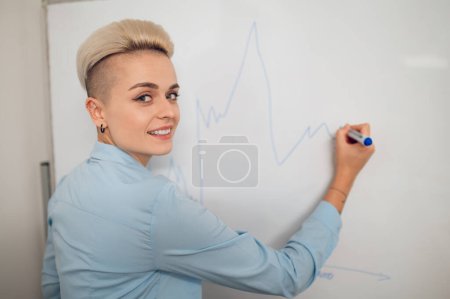 Photo for Smiling female corporate worker drawing the income graph with the marker on the whiteboard and looking back - Royalty Free Image