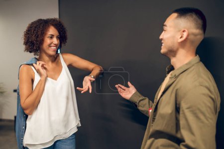 Foto de Sonriendo atractiva empleada de la compañía afroamericana hablando con un joven asiático en el pasillo - Imagen libre de derechos