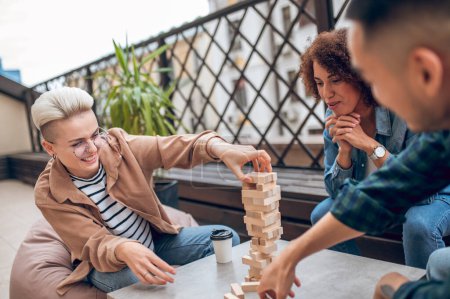 Photo for Smiling cheerful young woman placing the wooden blocks on top of the Jenga tower in the presence of her friends - Royalty Free Image