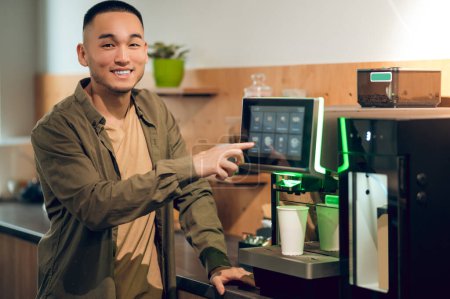 Photo for Waist-up portrait of a smiling cheerful young man tapping on the coffee machine touch screen and choosing the drink type - Royalty Free Image