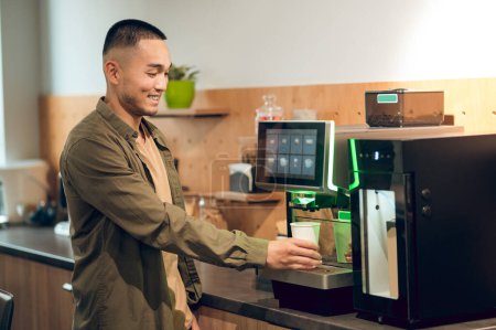 Foto de Sonriendo satisfecho hombre asiático poner una taza de papel desechable en la bandeja de goteo de la máquina de café - Imagen libre de derechos