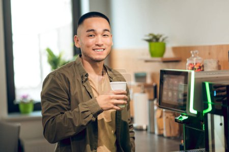 Foto de Retrato en la cintura de un empleado sonriente de la compañía parado en la cocina de la oficina y sosteniendo una bebida en la taza de papel - Imagen libre de derechos