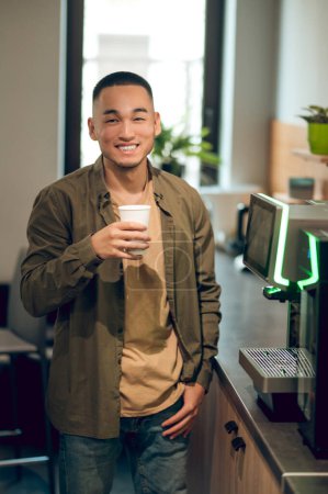 Foto de Alegre empleado de la compañía asiática sosteniendo una bebida en una taza de papel desechable en la mano y sonriendo a alguien - Imagen libre de derechos