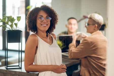 Téléchargez les photos : Travailleur d'entreprise souriant en lunettes appuyé sur le comptoir du bar et posant pour la caméra avec des collègues en arrière-plan - en image libre de droit