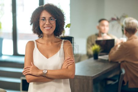 Téléchargez les photos : Joyeuse travailleuse d'entreprise en lunettes posant pour la caméra avec des collègues assis au comptoir du bar - en image libre de droit