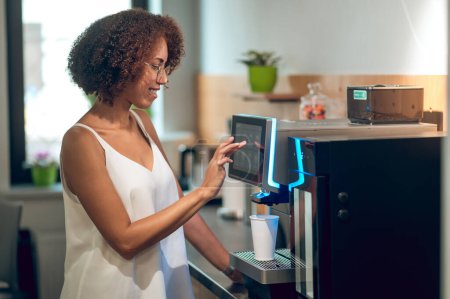 Foto de Sonriente mujer contenta tocando en la pantalla táctil de la máquina de café y elegir el tipo de bebida - Imagen libre de derechos