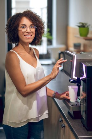Foto de Alegre empleado de oficina colocando la taza de papel en la bandeja de goteo y tocando en la pantalla táctil de la máquina de café - Imagen libre de derechos