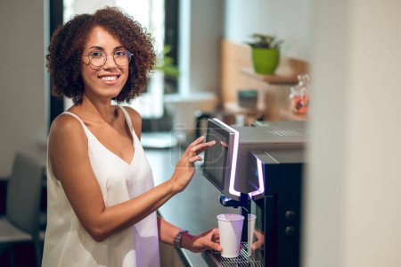 Foto de Trabajador de oficina sonriente poniendo una taza de papel desechable en la bandeja de goteo mientras golpea en la pantalla táctil de la máquina de café - Imagen libre de derechos