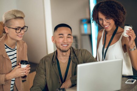 Téléchargez les photos : Travailleur d'entreprise heureux souriant assis à l'ordinateur portable en compagnie de ses collègues féminines - en image libre de droit