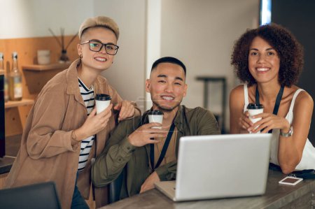 Foto de Sonriente gerente feliz sentado en el portátil rodeado de sus alegres colegas femeninos durante el descanso del café - Imagen libre de derechos
