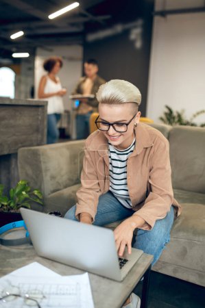 Téléchargez les photos : Une employée d'entreprise souriante portant des lunettes assise sur le canapé et travaillant sur son ordinateur portable - en image libre de droit
