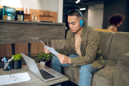 Photo for Serious focused corporate employee in wireless headphones seated on the sofa looking through documents in his hands - Royalty Free Image