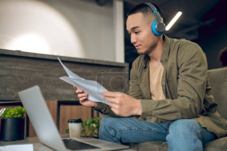 Photo for Serious concentrated young man in wireless headphones seated at the laptop looking through business documentation - Royalty Free Image