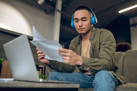 Téléchargez les photos : Travailleur d'entreprise dans les écouteurs sans fil assis à l'ordinateur portable tout en lisant des documents d'affaires dans ses mains - en image libre de droit