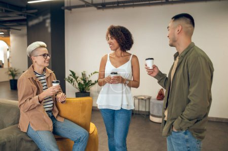 Photo for Group of three young joyful multinational corporate workers having a conversation during the coffee break - Royalty Free Image