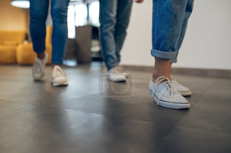 Foto de Foto recortada de piernas de tres personas en jeans y zapatillas de deporte de pie en el suelo - Imagen libre de derechos