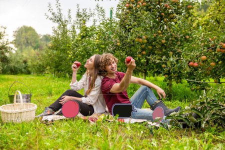 Foto de Feliz pareja romántica con manzanas en sus manos sentadas en la hierba entre los árboles frutales - Imagen libre de derechos