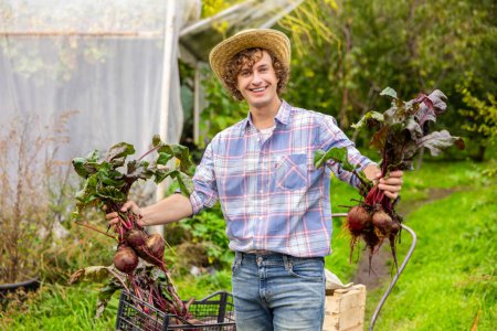 Photo for Smiling happy farm worker demonstrating young fresh organic beetroots with tops and long roots before the camera - Royalty Free Image