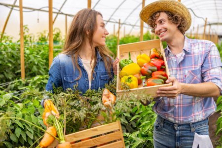 Téléchargez les photos : Heureuse agricultrice et sa joyeuse collègue tenant des caisses en bois avec les légumes récoltés et se souriant - en image libre de droit
