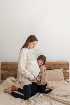 Foto de New life. Little girl looking interested while listening to babys moves in her moms belly - Imagen libre de derechos