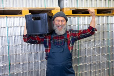 Foto de Cheerful factory worker standing beside beverage cans stacked on the pallets and holding a crate with bottles on his shoulder - Imagen libre de derechos