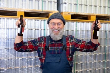 Photo for Cheerful brewery worker holding a pair of bottles in his hands while standing beside drink cans stacked on the pallets - Royalty Free Image