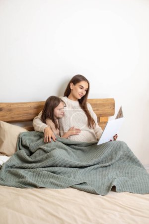 Photo for Mom and daughter. Pregnant young woman and a little girl on the bed looking peaceful - Royalty Free Image