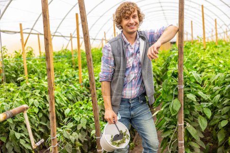 Téléchargez les photos : Cheerful agriculturist leaning against a wooden plant stake while holding a garden trowel and the bucket with pepper seedlings - en image libre de droit