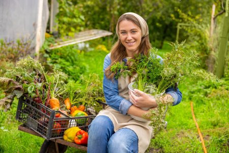 Téléchargez les photos : Smiling happy young female farm worker sitting on the cart loaded with fresh organic vegetables - en image libre de droit