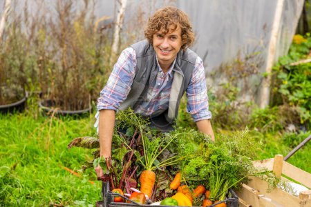 Téléchargez les photos : Smiling cheerful young Caucasian farmer placing a crate with fresh organic vegetables on the cart - en image libre de droit