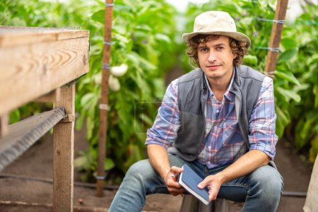 Foto de Calm vegetable grower holding a notebook and pencil in his hands sitting on the overturned bucket in the greenhouse - Imagen libre de derechos