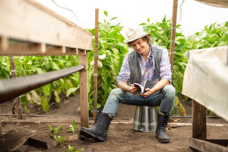 Photo for Smiling happy agronomist seated on the overturned bucket in the greenhouse leafing through his notebook - Royalty Free Image